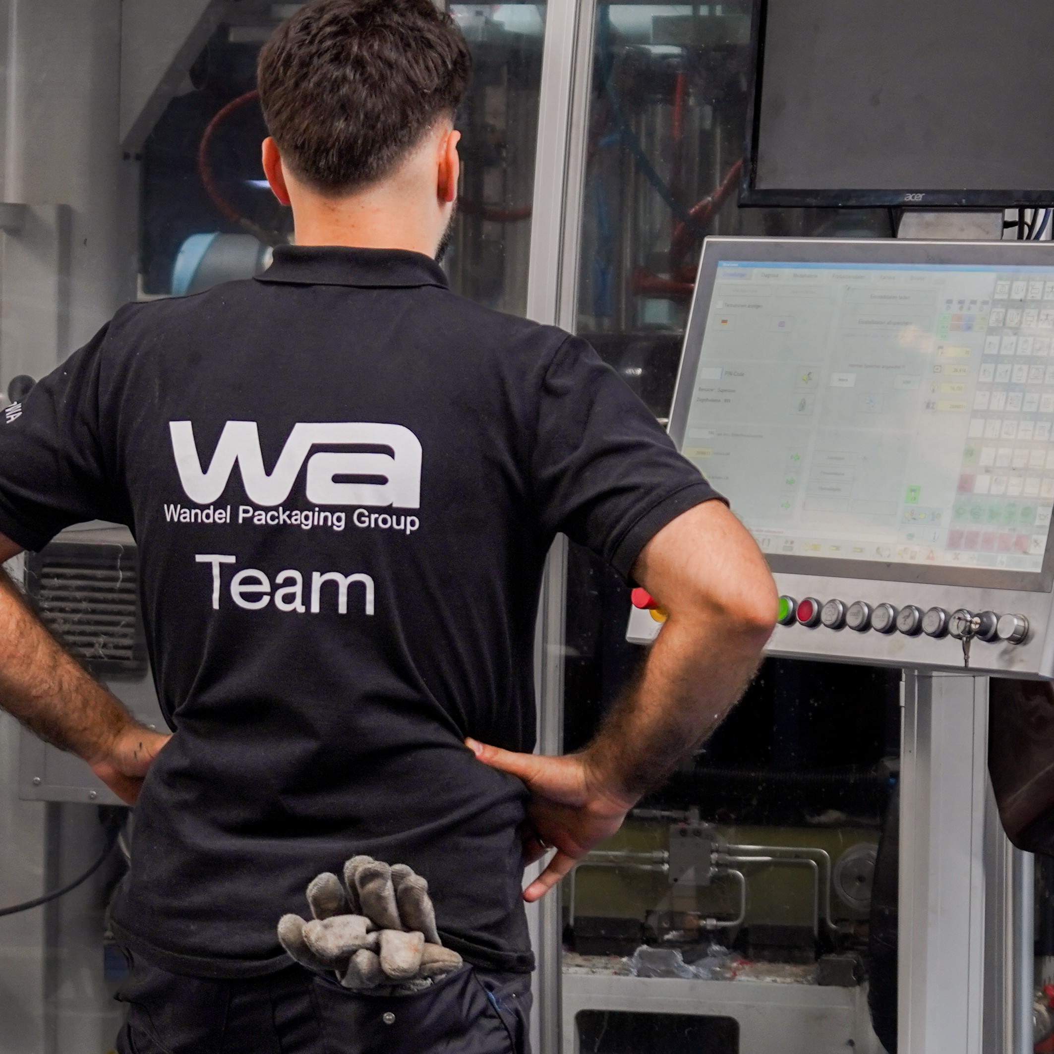 Maschinenbediener in schwarzem T-Shirt steht mit dem Rücken zum Fotograf vor einer Maschine